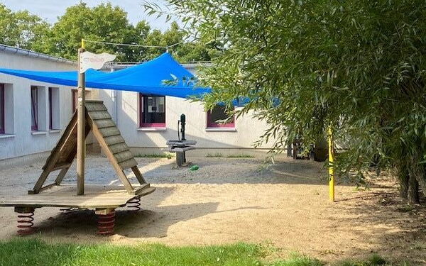 Kleiner Spielplatz vor einem Gebäude. Ein blaues Sonnensegel ist darüber gespannt und sorgt für Schatten.