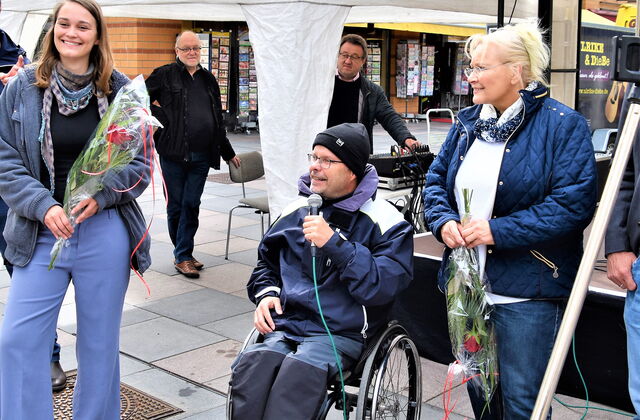 Zwei Frauen mit Blumen in den Händen. Zwischen ihnen ein Rollstuhlfahrer der in ein Mikro spricht. Dahinter ist ein weißer Pavillon. Der Rollstuhlfahrer trägt eine schwarze Mütze, Brille, Regenjacke. Er hat einen Aktivrollstuhl.