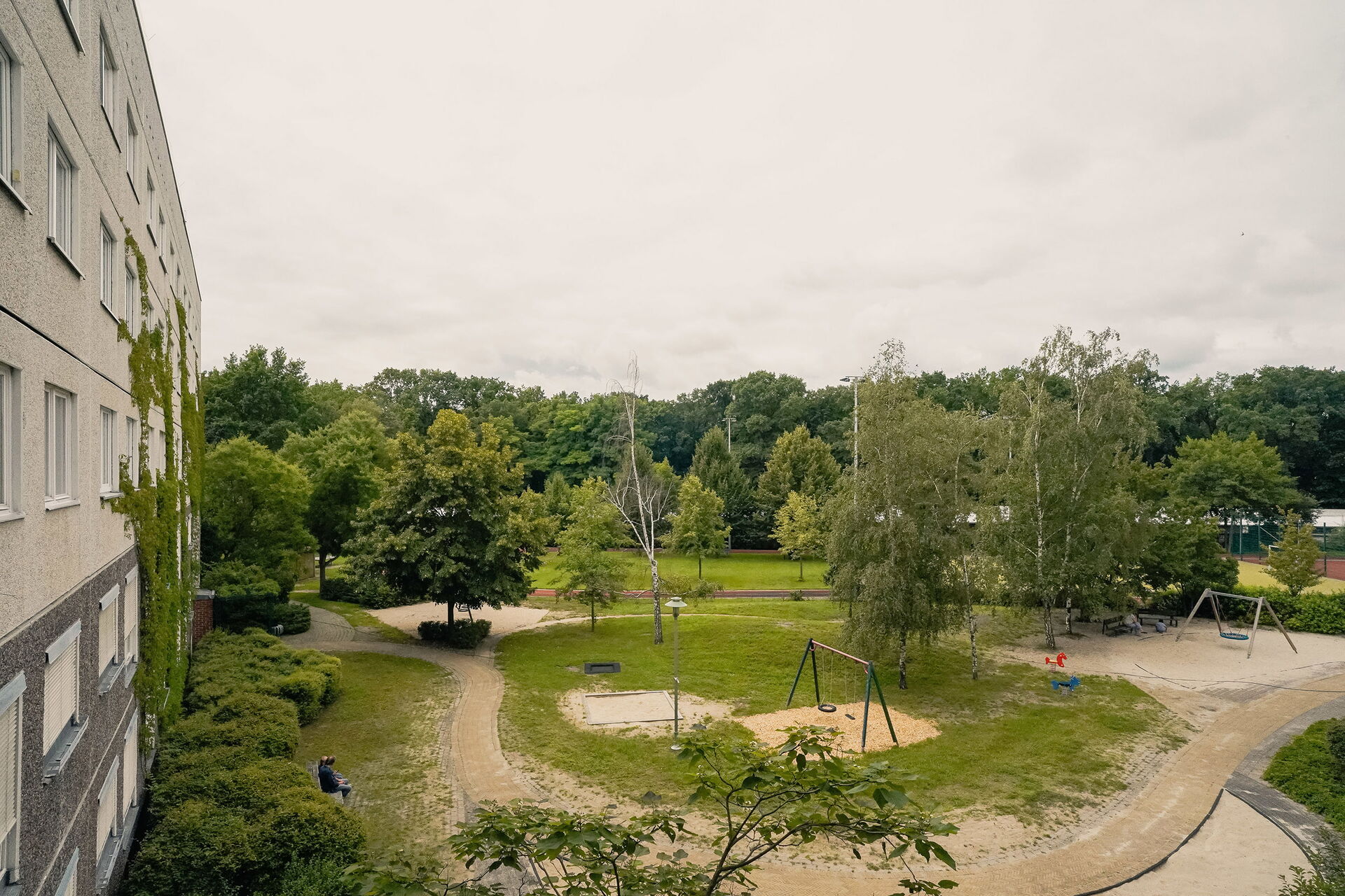 Blick aus einem oberen Fenster nach draußen auf einen Spielplatz. Sandkasten, Schaukeln, Wiese, ein paar Bäume.
