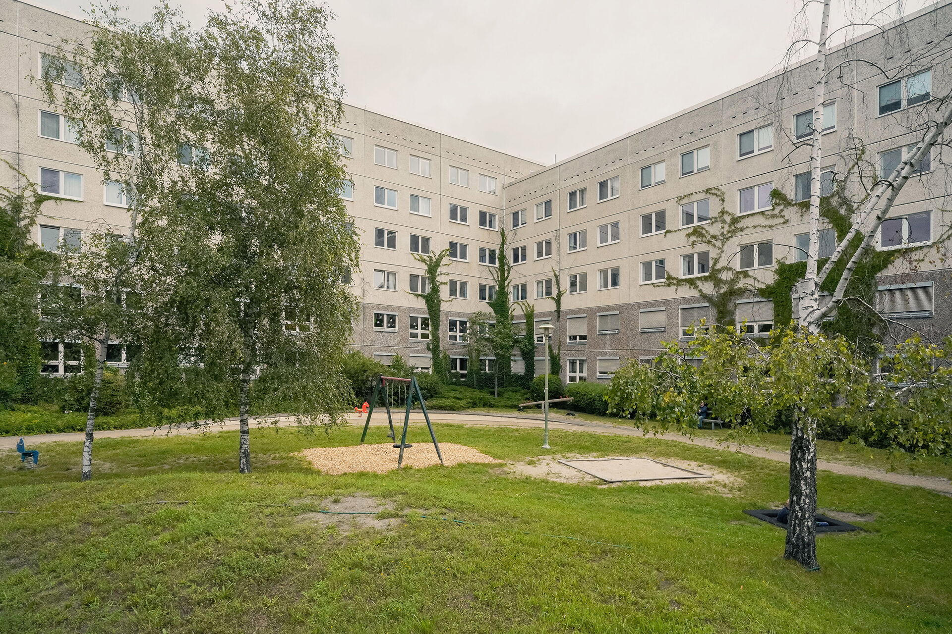 Draußen. Spielplatz, Schaukel, Wippe, Wiese, ein paar Bäume. Grauer Gebäudekomplex an einigen Stellen mit Efeu bewachsen.