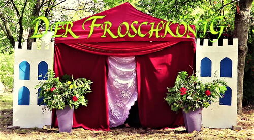 Draußen in der Natur. Ein rotes samtiges Märchenzeit, mit rosa Vorhang vor dem Eingang. Vor dem Zelt stehen links und rechts zwei Blumenkübeln. Schlossmauern aus Pappe sind links und rechts am Zelt.