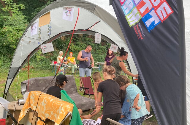 Großes Sonnensegel. Darunter eine Sitzecke aus Paletten und Decken.  Eine  Beachflag bedruckt mit dem Loge der AWO JUNGE PLATTFORM.