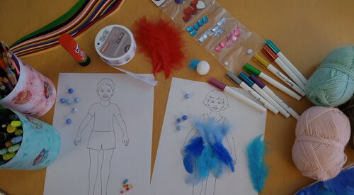 Tisch von oben mit Mal- und Bastelutensilien. Zwei Bilder nebeneinander, Junge und Mädchen. Auf dem Bild des Mädchens sind blaue Federn geklebt. Stifte, Schere, Wollknäuel, blaue Perlen, Prittstift