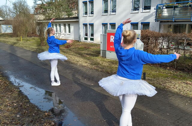 Draußen vor einem Gebäude. Pützen, ein Weg. Zwei weiße Ballett Tänzerinnen. Sie haben blonde lange haare, blaue Pullover, weißes Tutu, weiße Leggings und Turnschuhe an und tanzen.
