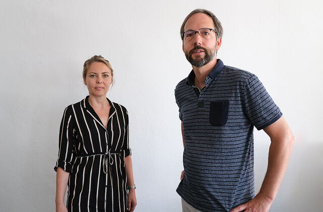 Zwei Personen. Frau und Mann vor einer weißen Wand. Frau ist blond und trägt eine schwarze lange Bluse mit weißen Längsstreifen. Mann mit Brille, Vollbart und blauem Poloshirt.