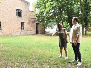 Draußen, ein altes Gebäude, Wiese und ein paar Bäume. Auf der Wiese im Vordergrund stehen eine Frau im Sommerkleid mit langen dunklen Haaren und ein Mann in T-Shirt, offenes Hemd darüber und langer Hose. Er sieht zum Gebäude.