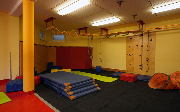 Turnhalle. 5 blaue Matten sind übereinander gestapelt. Eine Kletterwand mit Tritten. Kletterparcours an der Decke befestigt mit Stangen und unterschiedlichen Griffen.