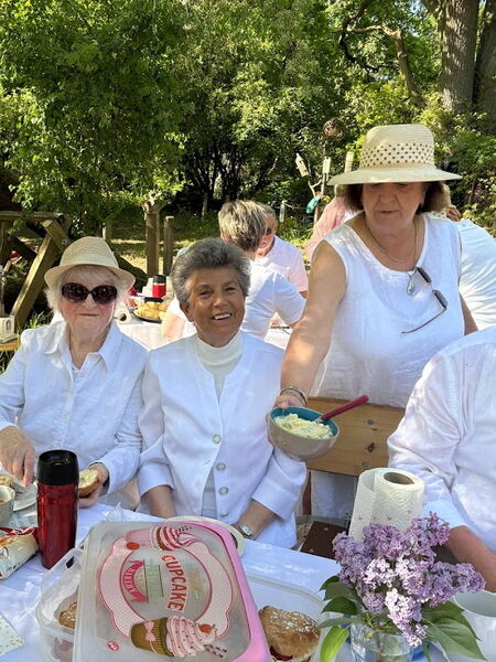 Frauen in weißer Kleidung und mit hellen Sommerhüten an einem Picknicktisch.