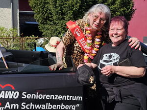 2 Frauen im schwarzen AWO Caprio. Frau mit grauen Haaren, roter Schärpe und Blumenkranz umarmt die andere Frau die am Auto lehnt.