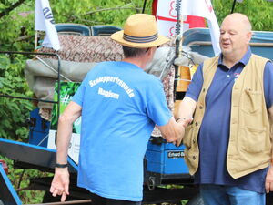 2 Männer begrüßen sich mit Handschlag vor einem blauen Trecker mit Anhänger.