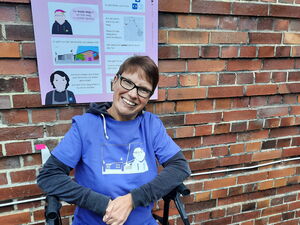 Ein Frau in T-Shirt und Jeans steht vor einer bunten Schrifttafel, die hinter ihr an einer Backsteinmauer angebracht ist.