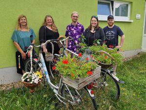 3 Frauen und zwei Männer stehen vor einer grünen Hauswand. Vor ihnen stehen zwei Fahrräder, auf den Gepäckträgern ist eine bepflanzte Holzkiste und ein bepflanter Korb mit Blumen. An einen Lenker hängt auch noch ein Körbchen mit Blumen.