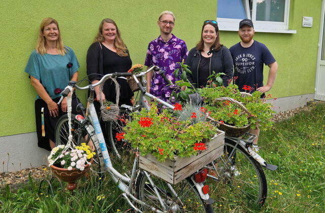 3 Frauen und zwei Männer stehen vor einer grünen Hauswand. Vor ihnen stehen zwei Fahrräder, auf den Gepäckträgern ist eine bepflanzte Holzkiste und ein bepflanter Korb mit Blumen. An einen Lenker hängt auch noch ein Körbchen mit Blumen.