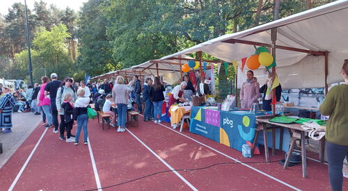 Bunte Marktstände, mit Luftballons und Wimpeln dekoriert. Viele Menschen von jung bis alt sehen sich die Stände an oder stehen beieinander.