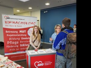 Ein AWO Stand auf einer Messe Mit Info- Material. Eine junge Frau steht hinter dem Stand und lächelt in die Kamera. Eine weitere Frau ist im Gespräch mit einem Mann.