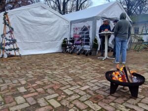 Draußen. 2 kleine Feuerschale im Vordergrund. Das große weiße Festzelt. Davor ein Stehttisch um den drei Personen stehen.