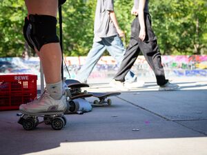 Draußen, eine Halfpipe, Skatbords. Beine mit Knieschützer, an den Füßen Rollschuhe. Zwei Jugendliche laufen vorbei.