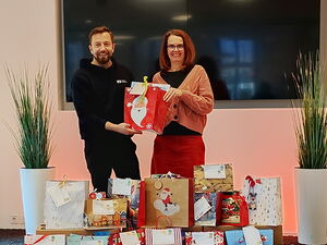 Gestapelte Weihnachtsgeschenktüten. Dahinter ein Mann und eine Frau die in die Kamera lächeln. Sie halten gemeinsam eine rote Weihnachtsmann-Geschenk-Tüte.