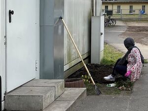 Draußen am Gebäude werden Blumen gepflanzt. Frau mit dunklem Kopftuch, hellem langärmligen Oberteil, rosa Gartenhandschuhe und weißen Turnschuhen sitzt auf dem Boden.