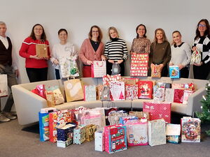 Ein Mann und 8 Frauen stehen hinter einer hellen Couch. Auf und vor der Couch viele Weihnachtsgeschenktüten. Rechts ein geschmückter Tannenbaum.