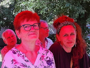 5 Personen sitzen an einer Biertischgarnitur unter einem roten Sonnenschirm. 2 Frauen im Vordergrund lächeln in die Kamera.