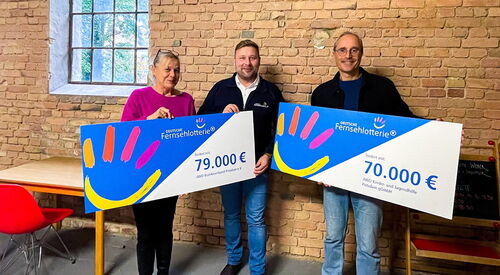 Eine Frau und zwei Männer halten die großen Schecks der Fernsehlotterie hoch und lächeln in die Kamera. Sie stehen in einem Raum vor einer Steinwand.