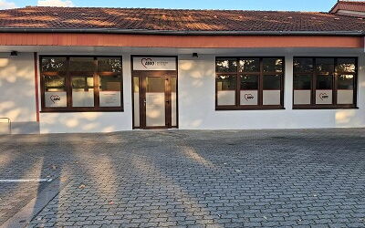 gepflasterte Parkfläche mit grauen Steinen, im Hintergrund ein Gebäude mit einer weißen Fassade, einem roten Dach, mit einer Glastür mittig, links von der Glastür eine Fensterglasfront, rechts der Glastür zwei Fensterglasfronten