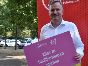 Portrait: Mann mit grauen kurzen Haaren. Er trägt ein weißes langärmliges Hemd und eine helle Hose. Er hält ein pinkes Schild vor sich. Text: Kitas zu Familienzentren machen.