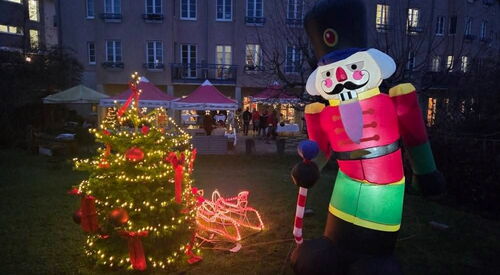 Im Vordergrund: kleiner geschmückter und beleuchteter Weihnachtsbaum, kleiner Schlitten und große aufblasbare Nussknackerfigur. Dahinter Pavillons und ein großes Gebäude. Es ist dunkel.