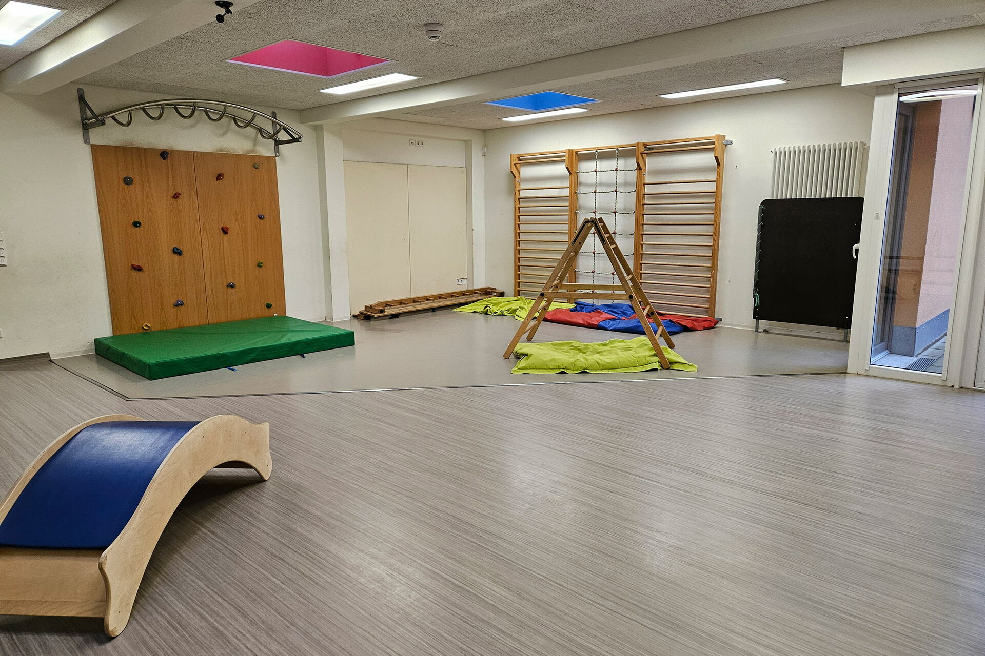 Raum mit Kletterelementen. Boulderwand, Sprossenwand und eine kleine Leiter mit schmalen Sprossen.