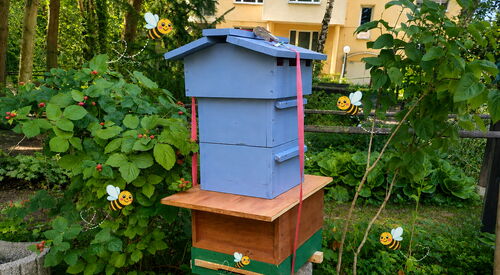 Aus Holz gebautes Haus mit mehreren Stockwerken für Bienen.   Einige virtuelle Bienen mit Gesichtern schwirren um das Häuschen.