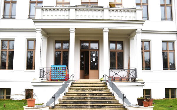 Eine Treppe führt hinauf zu der Eingangstür eines Gebäudes mit weißer Fassade und vier prächtigen Säulen. Die Eingangstür ist geöffnet.