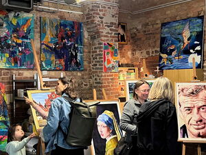 Menschen in einem Atelier, die sich Kunst Bilder, Gemälde ansehen. Es sind abstrakte Bilder aber auch Portraits darunter. Eine Frau im gelben Gewand und das Gesicht eines älteren Mannes mit Falten.