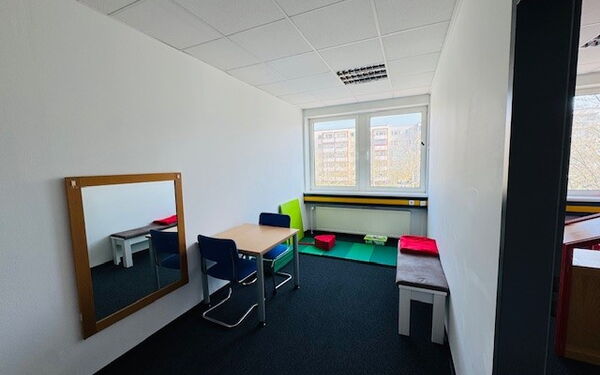 Ein sparsam ausgestatteter Raum mit einem Tisch und zwei Stühlen. An der Wand häng ein Spiegel, gegenüber steht eine Sitzbank. Unter dem Fenster gegenüber der Tür liegt eine Matte auf dem Boden. Darauf liegen ein Sitzkissen sowie ein Polster an der Wand.