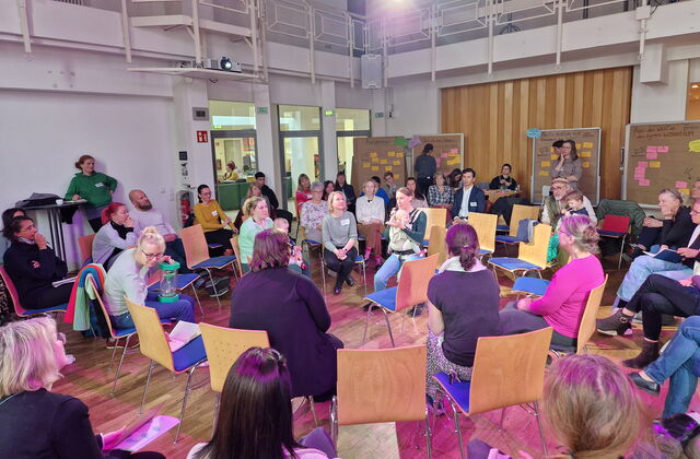 Großer Raum, Gesprächsrunde Menschen sitzen in Stuhlkreisen. Hinten im Raum sind 4 Flipcharts an denen farbige Moderationskarten angepinnt sind.