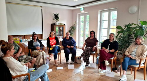 Teilnehmende mit Namenschildern vor sich auf dem Boden sitzen im Halbkreis auf Stühlen.