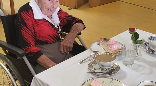 Seniorin im Rollstuhl am gedeckten Kaffeetisch. Nostalgisches Geschirr, ein großes Tortenstück und Kaffee stehen vor ihr.