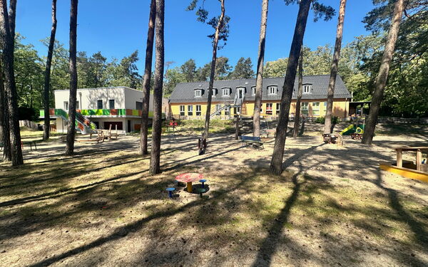 Draußen, Sonne scheint, Bäume sorgen für Schatten. Spielplatz. Das gelbe langgezogene Kitagebäude.