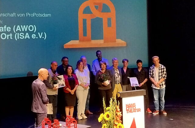 Auf der Bühne stehen 11 Menschen in einer Gruppe zusammen. Sie sind glücklich. Moderator Tim Jäger steht etwas abseits auch auf Bühne.