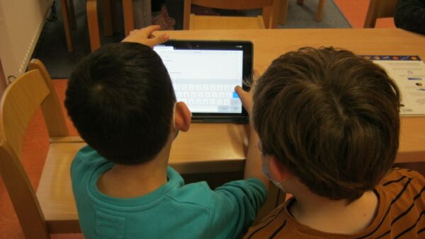 Zwei Kinder von hinten. Sie sitzen an einem kleinen Tisch. Ein Junge hält ein I-Pad in den Händen und drückt mit dem rechten Daumen auf ENTER. Das andere Kind guckt im dabei zu.