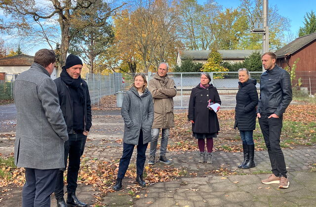 Herbst, Draußen, blauer Himmel, Schäfchenwolken. 7 Personen stehen draußen verteilt. Alle sehen zu Brain Nickholz.