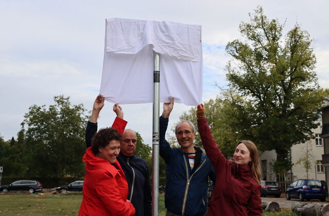 Ein Straßenschild mit einem weißen Tusch darüber. Von unten greifen 2 Frauen und 2 Männer nach dem Tusch. Die Enthüllung steht kurz bevor.