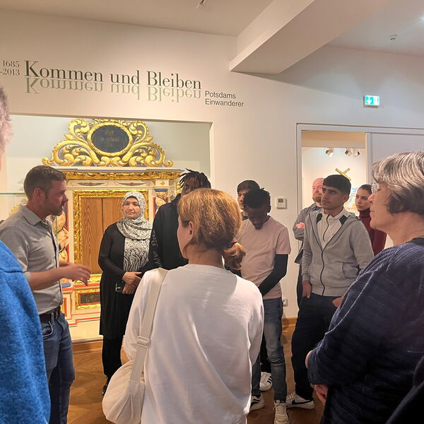 Menschen bei einer Führung durchs Museum. Sie stehen in einem Raum, Ein der Wand steht der Schriftzug: "1685 bis 2013 - Kommen und Bleiben - Potsdams Einwanderer"