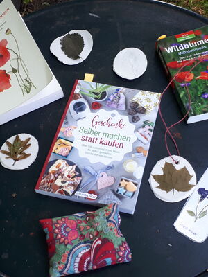 Auf einem runden schwarzen Tisch liegen Naturbücher und kleine Schalen mit Blüten und Blättern von Bäumen, in der Mitte das Buch: ""Geschenke selber machen statt kaufen"