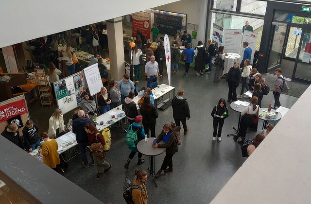 Ein Foyer von schrägoben. Stehtische und Info-Stände. Mehrere Menschen stehen an den Tischen und stehen im Raum herum.