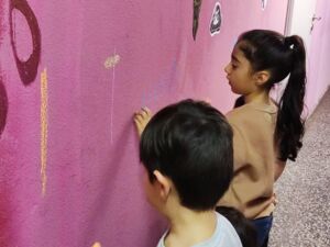 Ein Junge und ein Mädchen stehen an einer pinken Wand. Das Mädchen malt etwas darauf. Das Mädchen har lange schwarze Haare zu einem hohen Zopf gebunden, sie trägt einen hellbraunen Pullover.