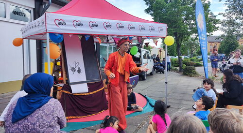 Ein farbenfroh gekleideter Mann mit Narrenmütze bespaßt Kinder und Erwachsene. Er trägt Orange-Rote Kleidung.