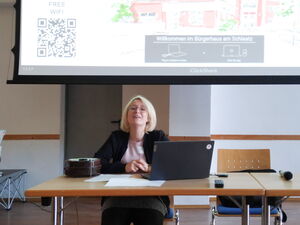 Blonde Frau mit Brille sitzt an einem Tisch mit Laptop. Begrüßung.