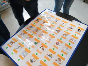 Auf einem Tisch liegt eine laminierte Tafel mit Symbolkarten und Bildern, ein Kommunikationsboard. Drei Personen stehen darum und schauen darauf.