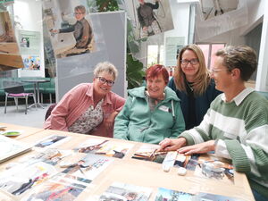 4 Frauen an einem Tisch, drei sehen und lächeln in der Kamera. Rechts sitzt eine frau und zeigt auf Fotos die auf dem Tisch liegen.